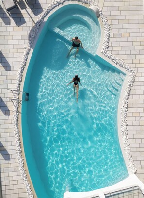 Une vue aérienne d’une piscine où l’on aperçoit deux personnes dans l’eau, le tout étant entouré d’une bordure de galets et d’une terrasse carrelée.