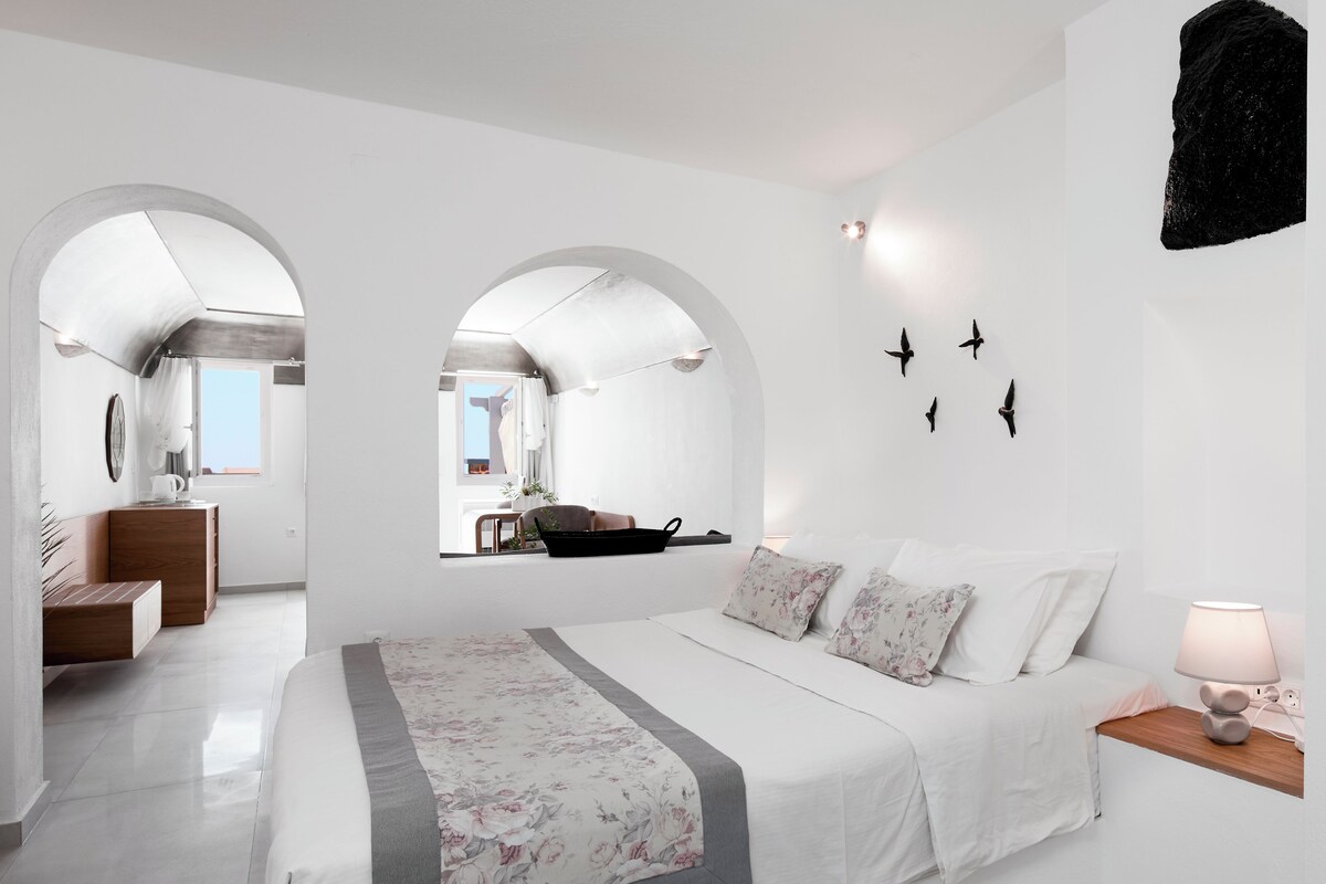 A modern bedroom with a large bed, a wooden nightstand, a desk, and a view through an arched window.