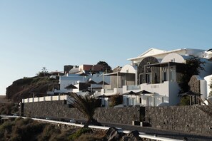 A coastal hotel with modern architecture, featuring multiple white buildings with curved roofs, a stone retaining wall, and a palm tree in the foreground. - Santorini Princess Spa Hotel (Santorini)