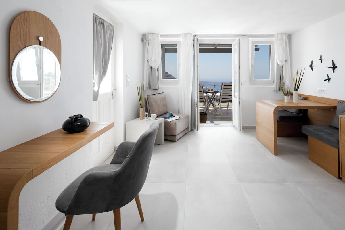 A modern, well-lit room with a wooden desk, a grey chair, a small sofa, and a view of the sea through open doors.