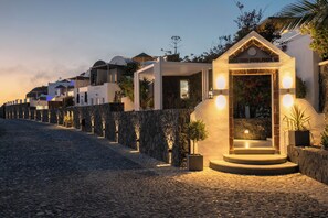 Una calle empedrada bordeada por edificios blancos modernos, un muro de piedra y una pequeña estructura iluminada con escalones que conducen a una puerta.