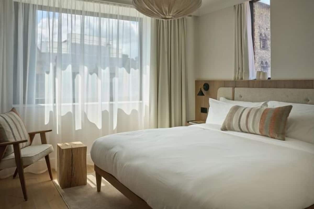 Hotellihuone, jossa on suuri sänky, tuoli, yöpöytä ja ikkuna, jossa on ohuet verhot.