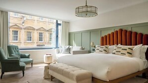 A hotel room with a large bed, a green armchair, a bench, and a view of a building through the window. - The Store (Oxford)