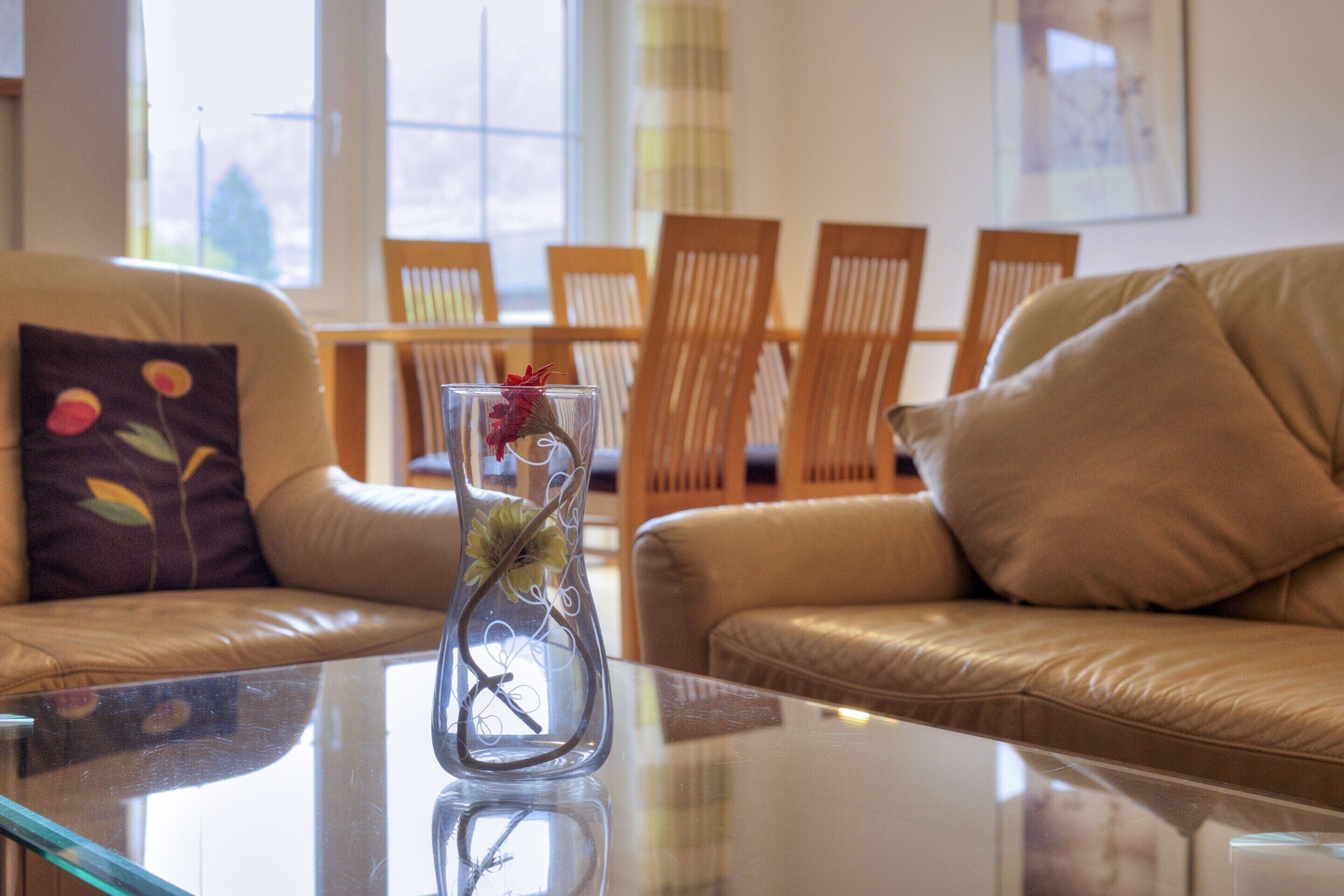 A living room with a glass coffee table, a vase with flowers, and a sofa with a cushion.