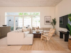 Een moderne woonkamer met een hoekbank, een salontafel, een rieten fauteuil en een aan de muur bevestigde televisie.