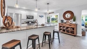 A modern kitchen with white cabinets, a large island with stools, pendant lighting, and a wooden cabinet with drawers. - Updated! Heated Pool! Near Beach! Grill, Parking, Outdoor Oasis! ~ Rosemary's Hacienda (Largo)