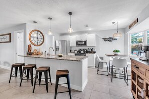 A modern kitchen with white cabinets, a central island with stools, pendant lighting, and a large wall clock.
