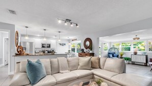 A modern living room with a beige sectional sofa, a coffee table with decorative items, and a kitchen in the background. - Updated! Heated Pool! Near Beach! Grill, Parking, Outdoor Oasis! ~ Rosemary's Hacienda (Largo)