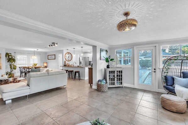 A spacious living area with a sofa, dining table, and chairs, a kitchen in the background, and a large window with a view.