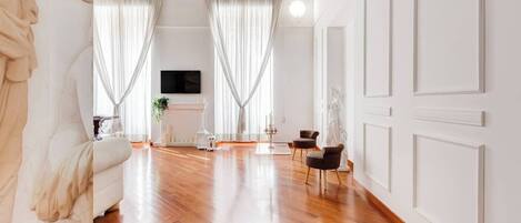 A spacious living room with wooden flooring, white walls, and large windows with sheer curtains. There is a fireplace, a television, and two chairs.