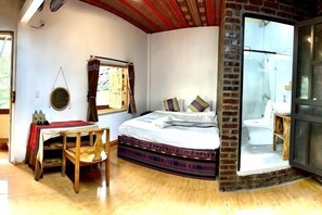 Une chambre à coucher avec un lit, une table et une chaise. Il y a une fenêtre avec des rideaux, une porte donnant sur une salle de bain et un mur en briques.