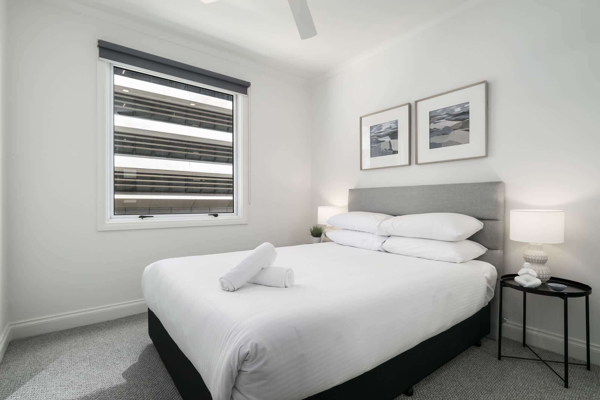 A neatly arranged bedroom with a bed, a bedside table with a lamp, a window with blinds, and two framed pictures on the wall.