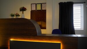A reception desk with a wooden finish and a built-in light. There are two potted plants on the desk, a bulletin board with framed notices, and a window with curtains. - Kavitha Stay Inn (Namakkal)