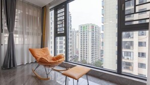A modern room with a large window, a rocking chair, a small table, and a view of high-rise buildings. - My Heart Returns · Zhangjiajie Zero-Pressure Luxury Dream Apartment (Zhangjiajie)