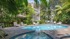 Una piscina con lettini e una zona coperta, immersa nel verde e affiancata da un edificio a più piani.