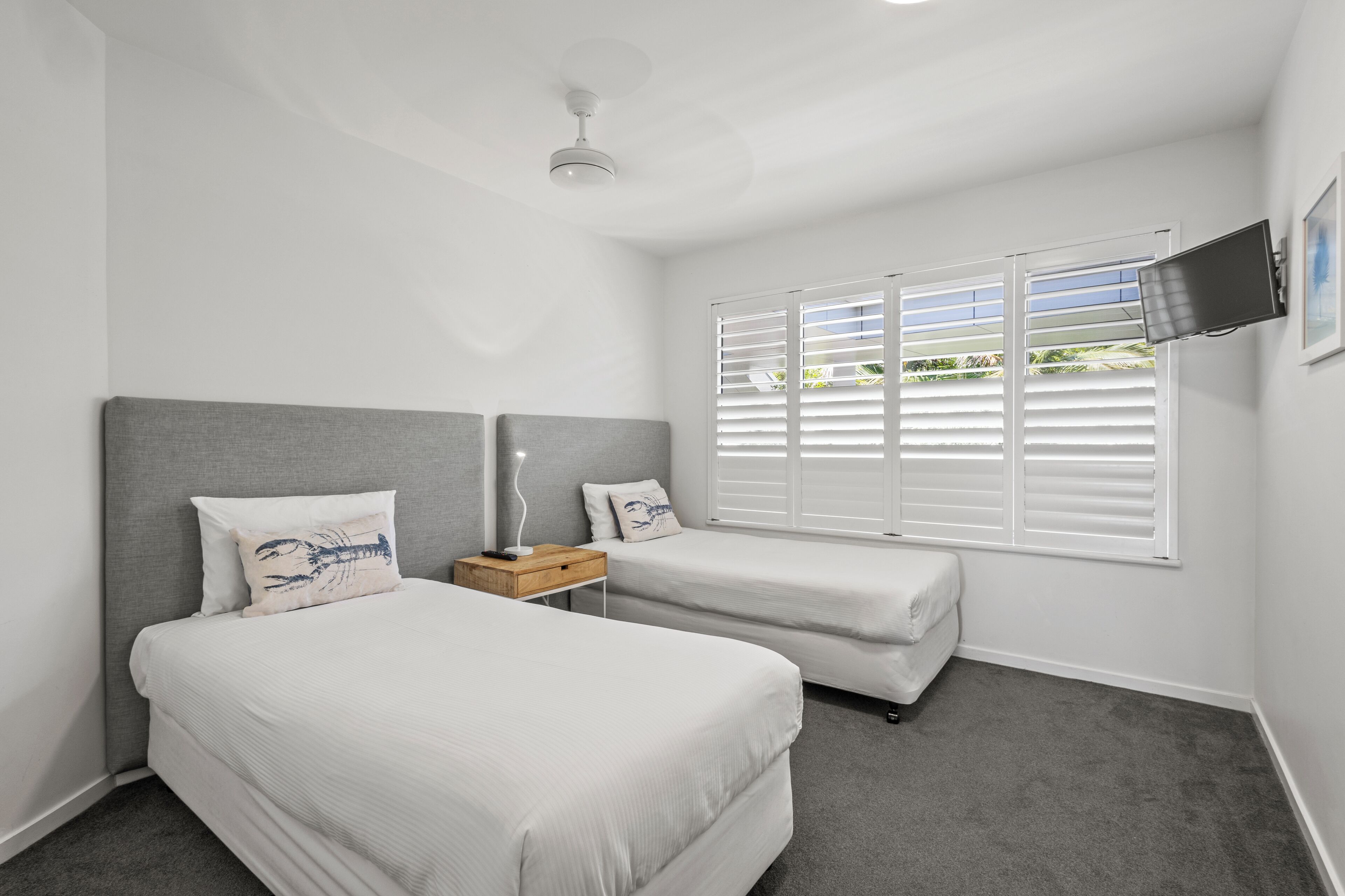 A bedroom with two beds, a ceiling fan, a TV mounted on the wall, and a window with blinds.