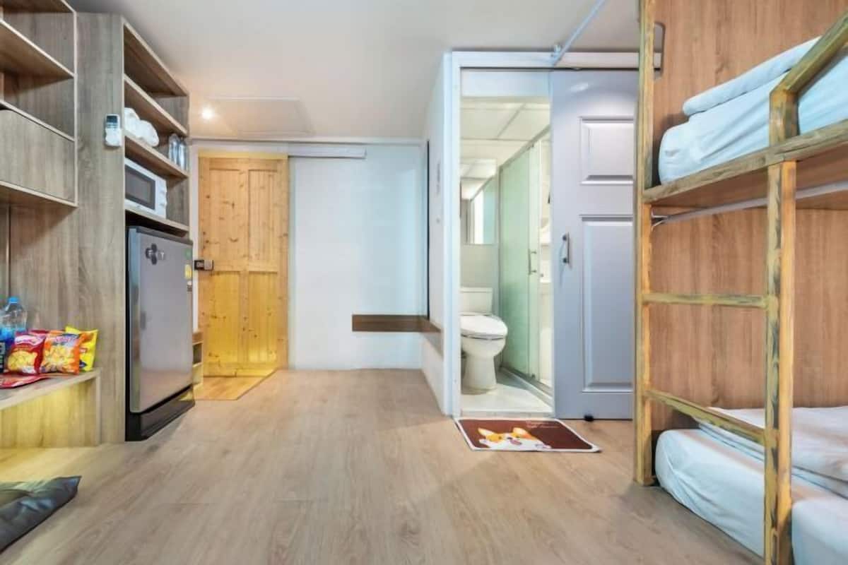 Ein kleines Zimmer mit Küchenzeile, Holzregalen, einem Etagenbett und einer Tür zu einem Badezimmer.