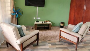 A living room with two beige armchairs, a wooden coffee table, a television mounted on the wall, a fan, and a drum. - Mew3 Apartments (Ndola)