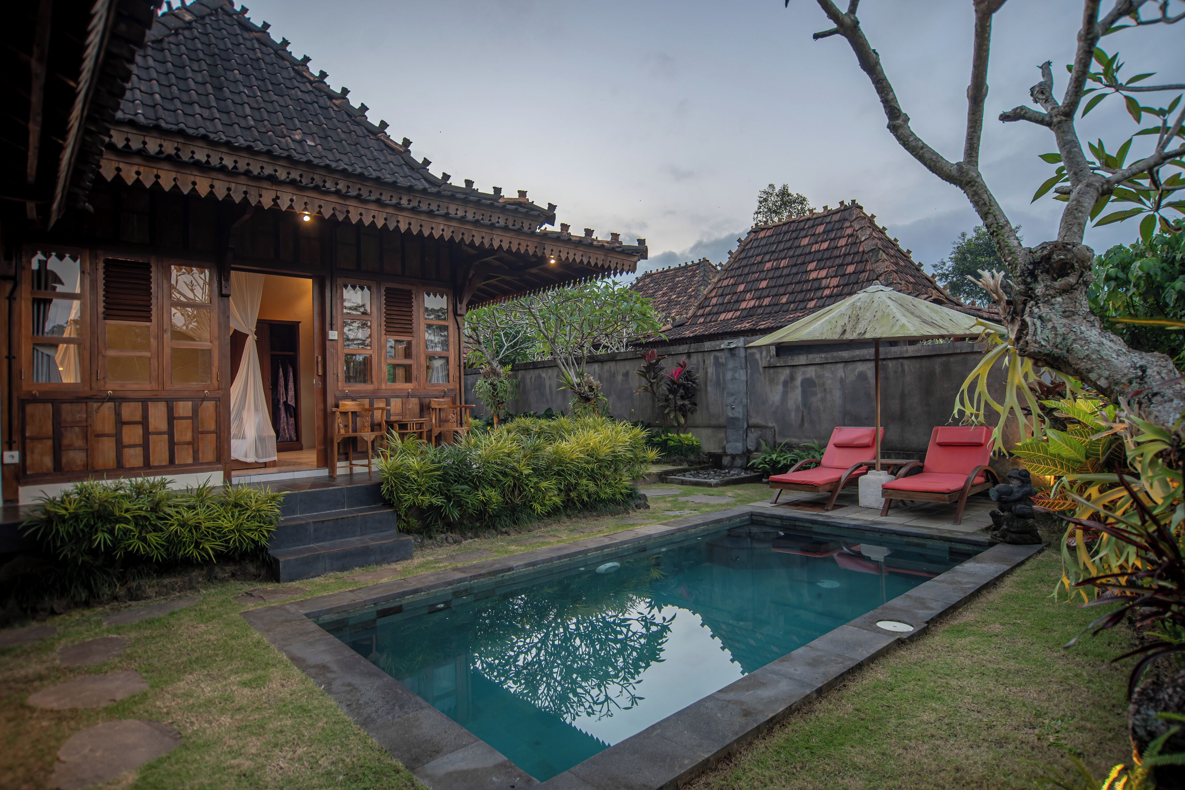A traditional wooden house with a swimming pool, red lounge chairs, and a stone pathway.