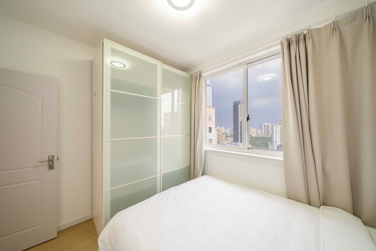 A bedroom with a bed, a wardrobe, a window with curtains, and a view of buildings outside.