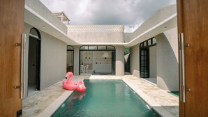 A modern pool area with a pink flamingo float, a covered seating area, and a view of the interior space. - SOOJIWA VILLA by Taman Bali (Canggu)