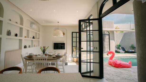A dining area with a round table and chairs, a hanging light fixture, and a view of a swimming pool with a pink flamingo float. - SOOJIWA VILLA by Taman Bali (Canggu)