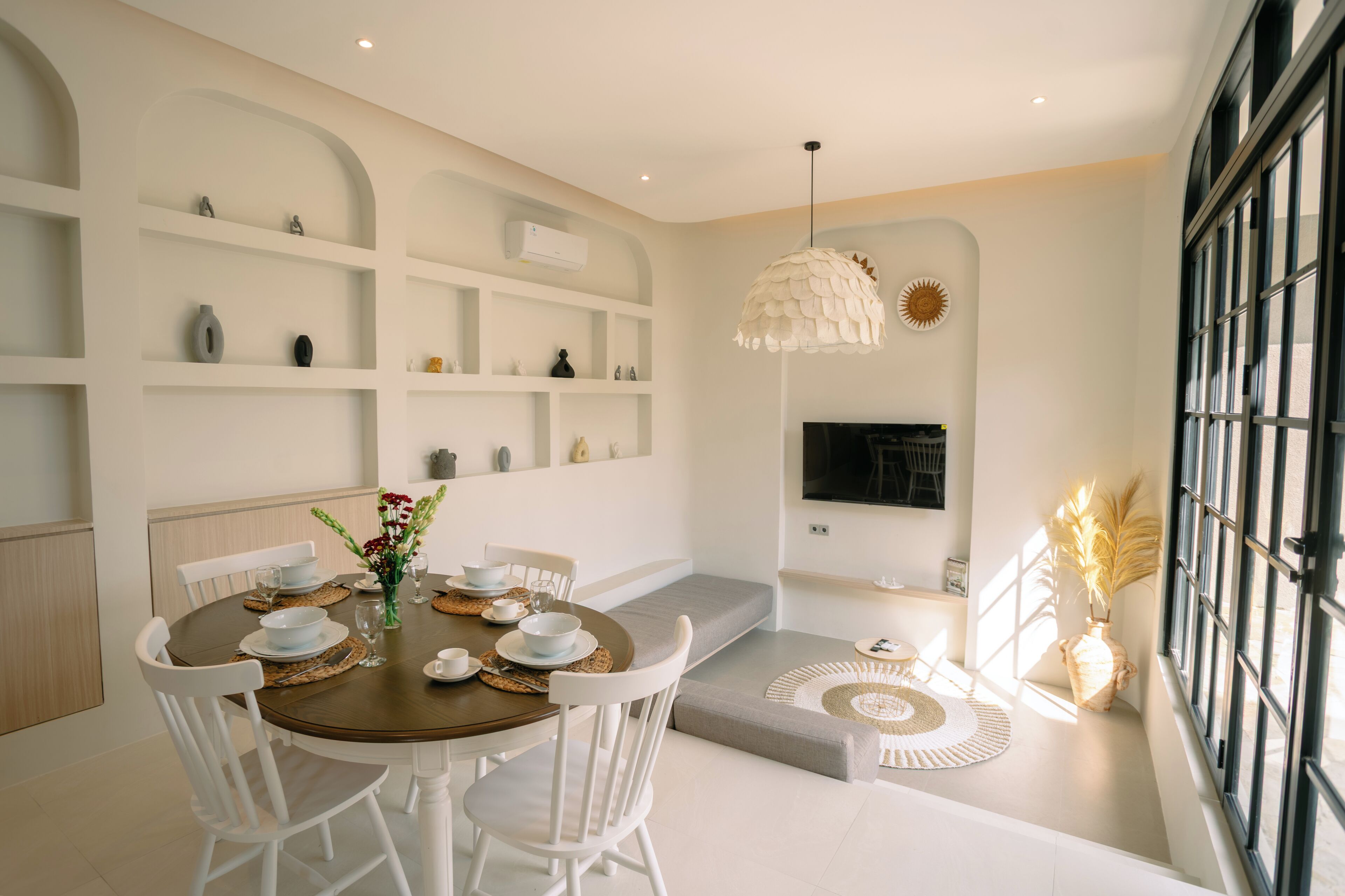 A dining area with a round table, white chairs, and a central vase with flowers. There are shelves with decorative items and a television on the wall.
