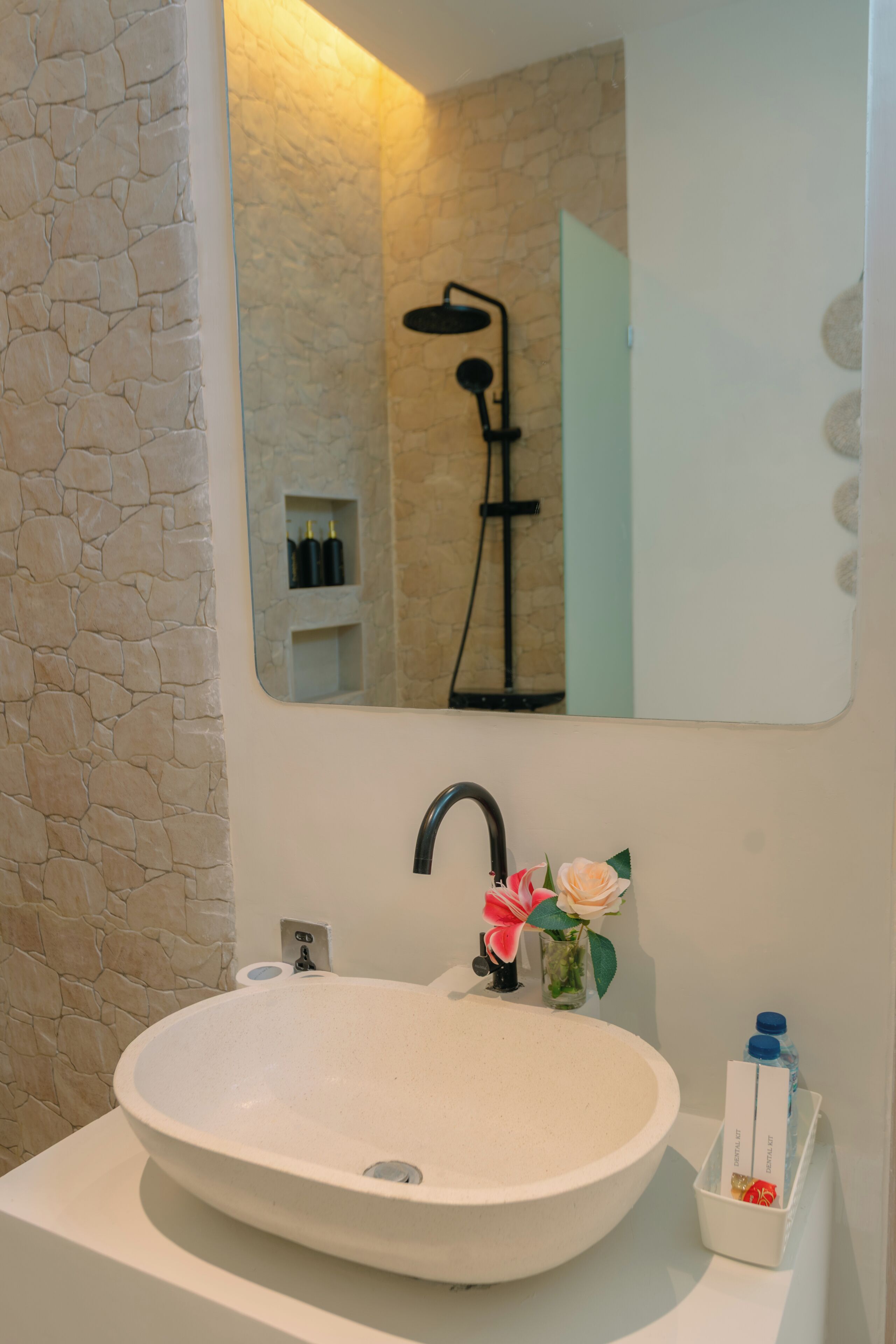 A bathroom with a stone wall, a round sink, a mirror, a shower, and toiletries.