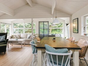Un vaste espace dédié à la salle à manger, avec une table et des chaises en bois, de grandes fenêtres offrant une vue sur la verdure, et un plafond blanc avec des poutres apparentes.