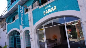 Un hotel con un cartello ben visibile che recita 'HOTEL DAMAR', una hall con ingresso adiacente a una vetrata e una zona esterna arredata con divano e tavolo.