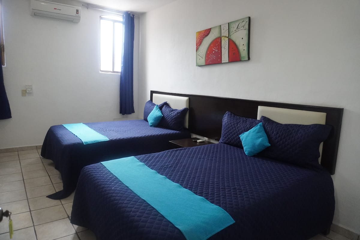 Une chambre d’hôtel avec deux lits, chacun doté d’une tête de lit et de draps bleus. On y trouve une fenêtre avec des rideaux, un climatiseur fixé au mur et une œuvre d’art décorative au-dessus des lits.