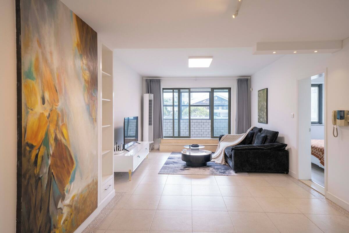 Un salon spacieux agrémenté d’un grand tableau abstrait, d’un canapé moderne, d’une table basse et d’une grande fenêtre donnant sur un immeuble.