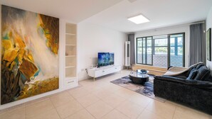 A modern living room with a large abstract painting, a flat-screen TV, a dark sofa, and a large window offering a city view. - Tyms Family 3BR 2BA APT Crystal Garden (Shanghai)