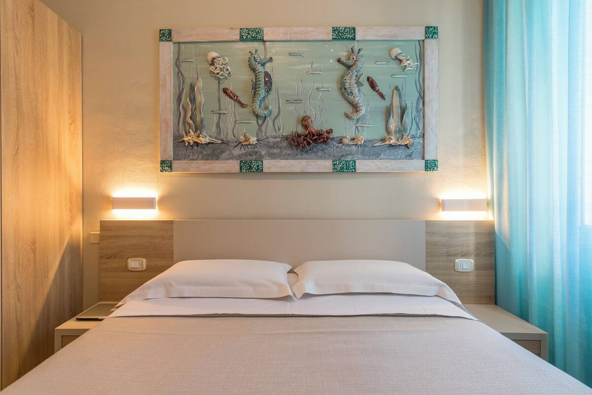 Habitación de hotel con cama, dos lámparas de noche, una obra de arte en la pared que representa vida marina y cabecero de madera.