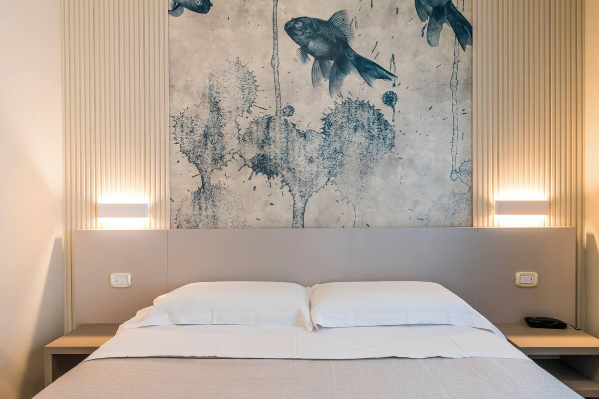 Una cama bien hecha con sábanas blancas en una habitación de hotel, una pared con un cuadro abstracto grande, y dos lámparas de noche.