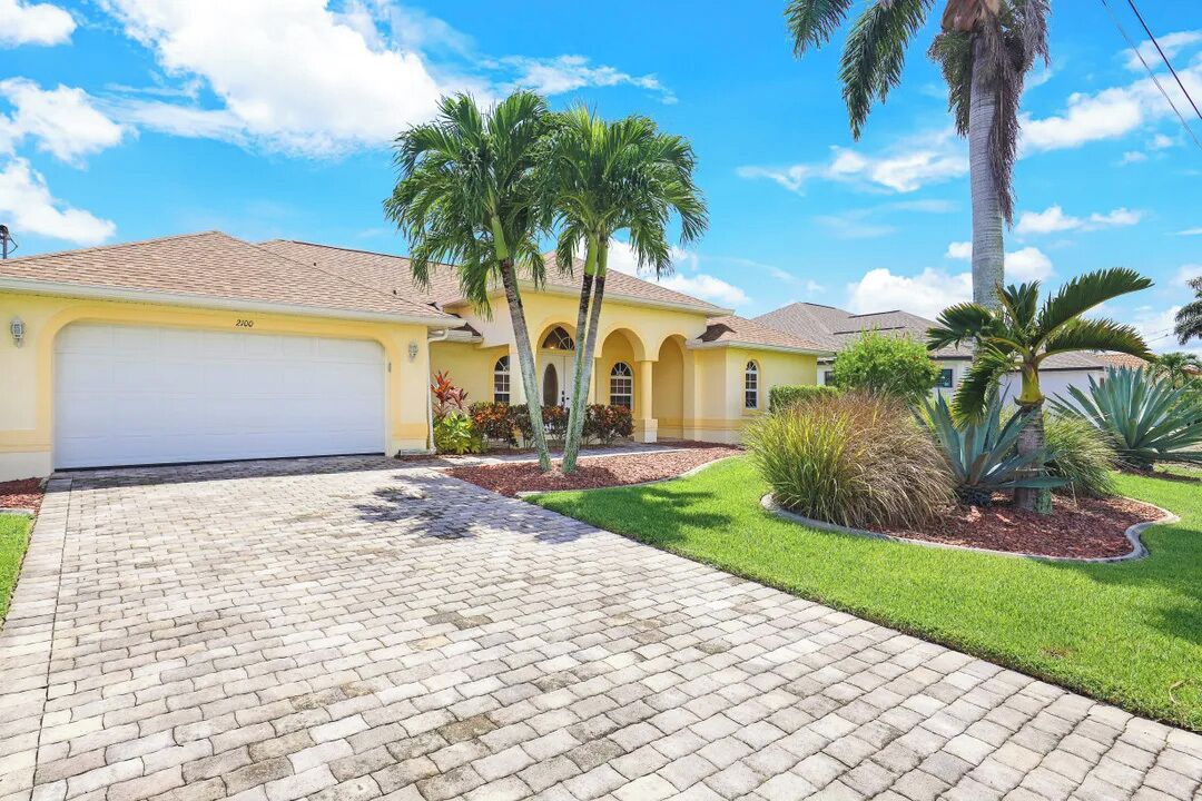 A yellow house with a garage, palm trees, and a paved driveway.