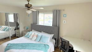 Ein Schlafzimmer mit einem Bett, einem Schreibtisch, einem Stuhl, einem Deckenventilator und einem Fenster mit Vorhängen.
