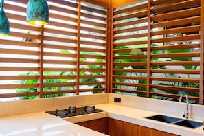 A kitchen with a built-in stove, a sink, and wooden blinds with a view of green foliage. - Ersanea Villas Seseh (Bali)
