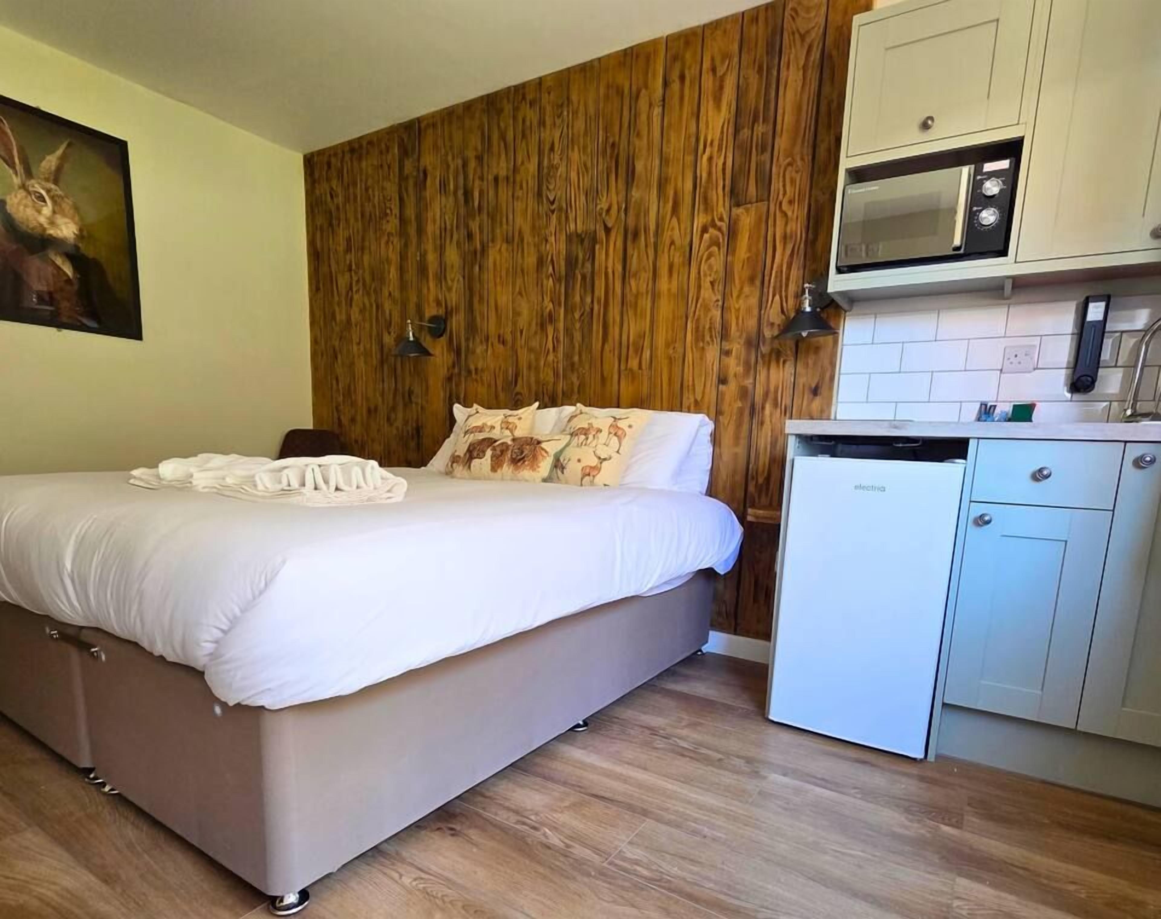 A bedroom with a wooden wall, a bed with white bedding, and a kitchenette with a microwave and a dishwasher.