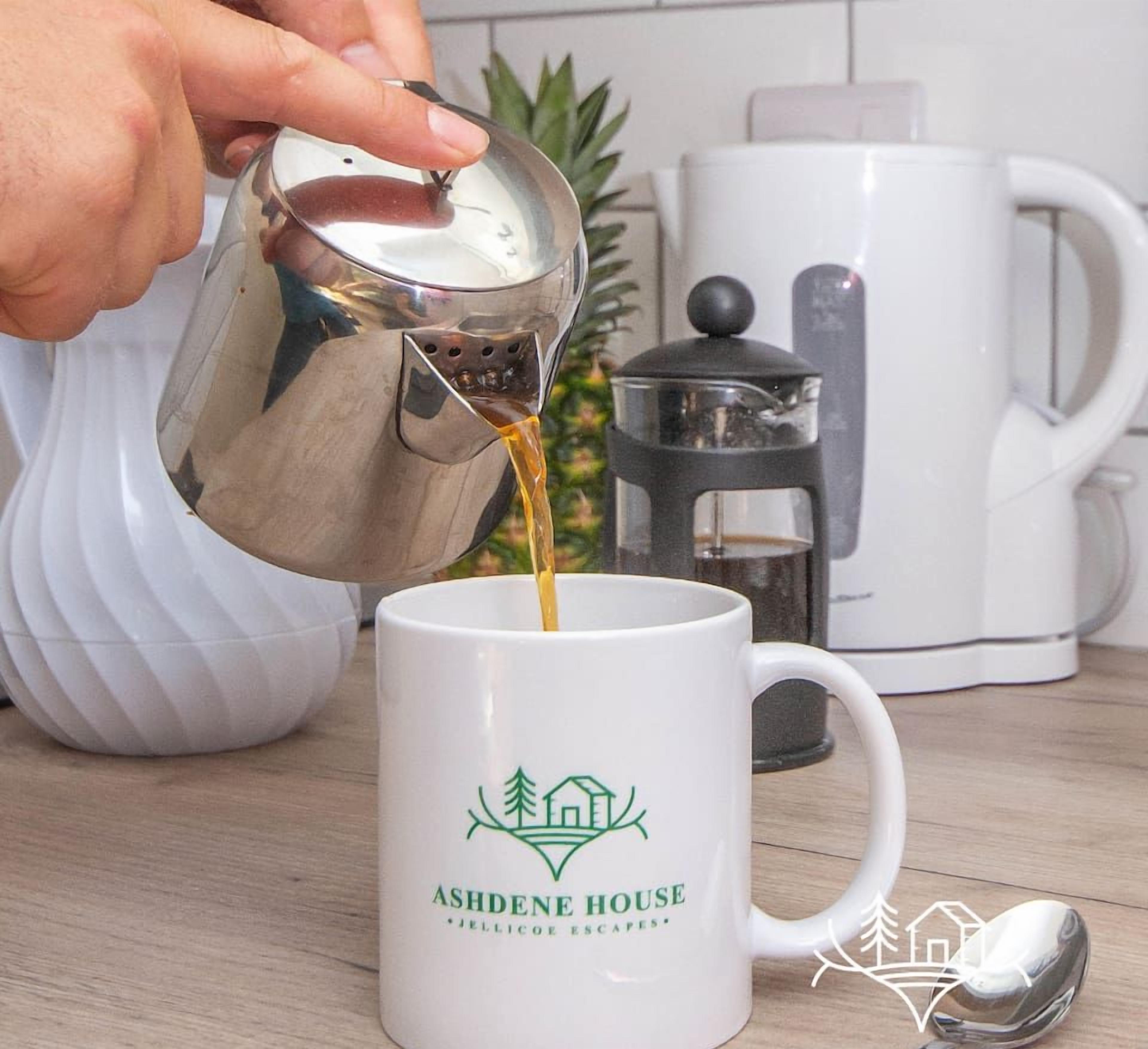 Une main verse du café dans une tasse ornée d’un logo et d’une inscription indiquant « ASHDENE HOUSE JELLICOE ESCAPES ».