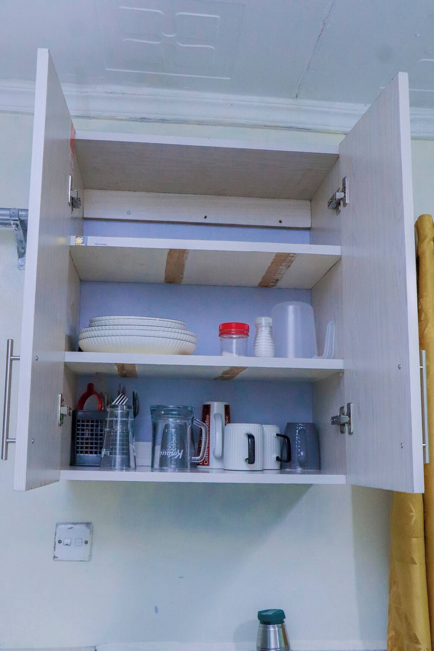 A kitchen cabinet with shelves containing plates, cups, a coffee maker, and a kettle.