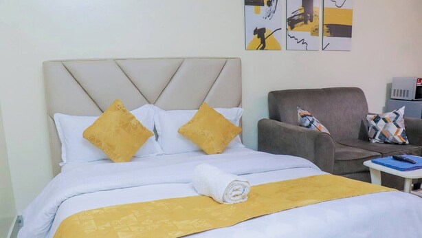 A neatly made bed with a white and yellow bedspread, two pillows, and a rolled towel. There is a gray armchair with a patterned cushion and a small table with a remote control.