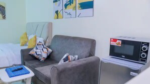 A compact hotel room with a grey sofa, a small white table with a remote control, and a microwave on a portable stand.