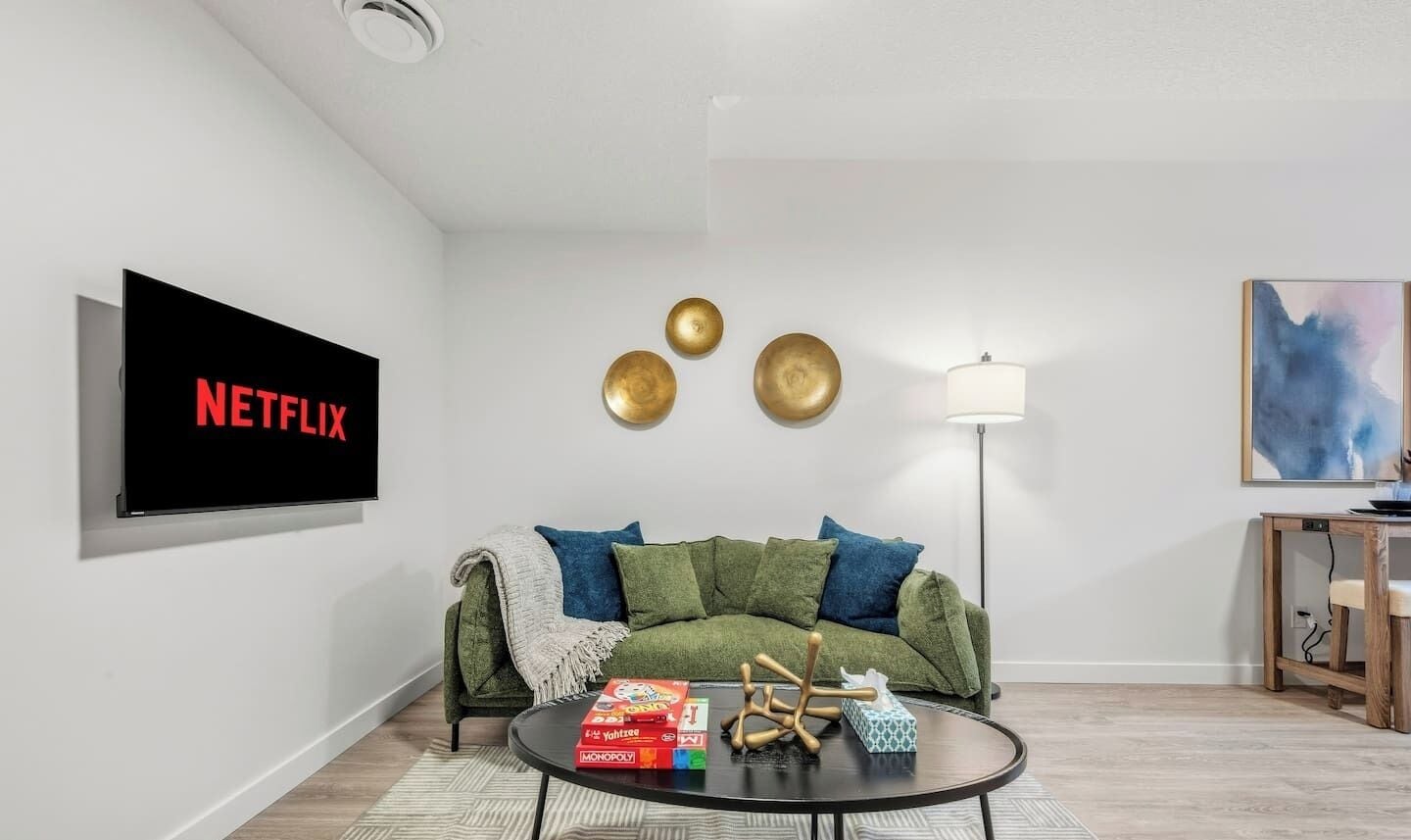 En moderne stue med en grøn sofa, et sofabord med brætspil, et fladskærms-tv der viser 'NETFLIX' og en lampe.