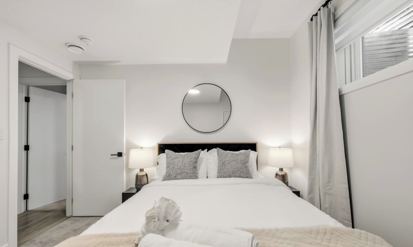 Una habitación de hotel con una cama grande, dos mesitas de noche con lámparas, un espejo redondo en la pared y una puerta que da a otra habitación.