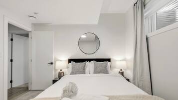 Una habitación de hotel con una cama grande, dos mesitas de noche con lámparas, un espejo redondo en la pared y una puerta que da a otra habitación.