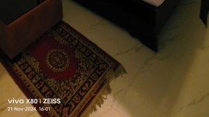 A bedroom with a bed, bedside tables, a ceiling fan, an air conditioning unit, a wall-mounted light, and a decorative rug. - Hotel Prince Palace (Ayodhya)