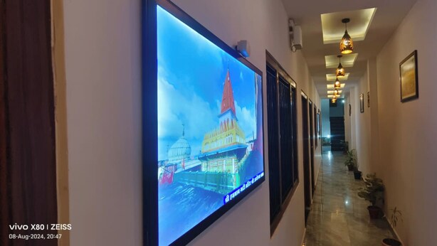 A hallway with a large digital display on the left, a ceiling with hanging lights, and a staircase in the background. - Hotel Prince Palace (Ayodhya)