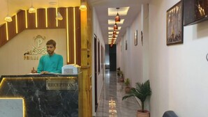 A reception desk with a marble top, a sign reading "RECEPTION," and a logo above it. There is a staircase to the left and a hallway with potted plants and framed pictures on the walls. - Hotel Prince Palace (Ayodhya)