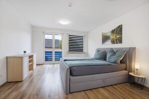 A bedroom with a bed, a desk, a window with blinds, and a painting on the wall.